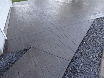 A textured concrete pathway with a stamped design, flanked by a bed of small stones, next to a structure with white siding.