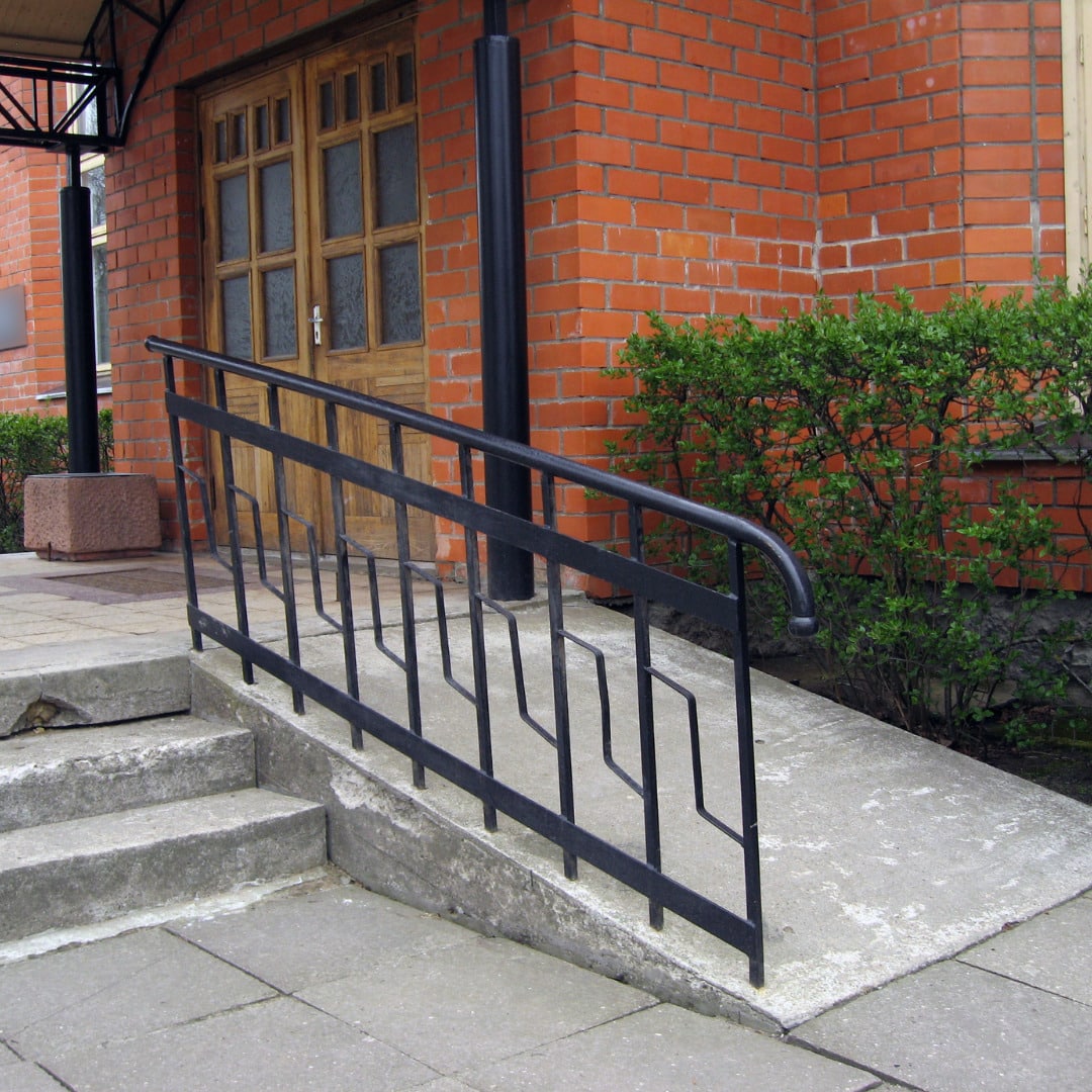 Black metal handrail beside concrete steps leading to a wooden door of a brick building with green shrubbery.