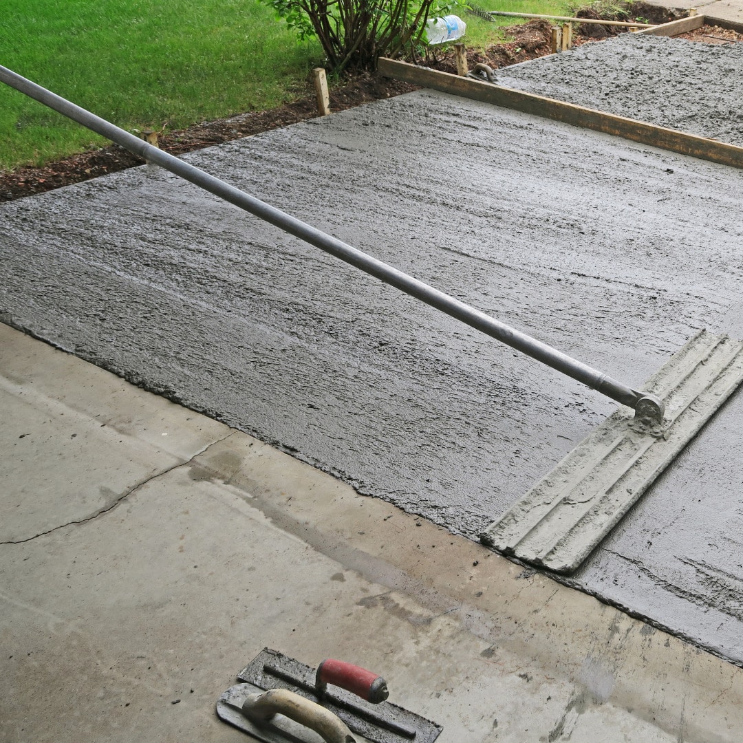 Fresh concrete being screeded with a long metal straightedge, ensuring a level surface with tools visible in the foreground.