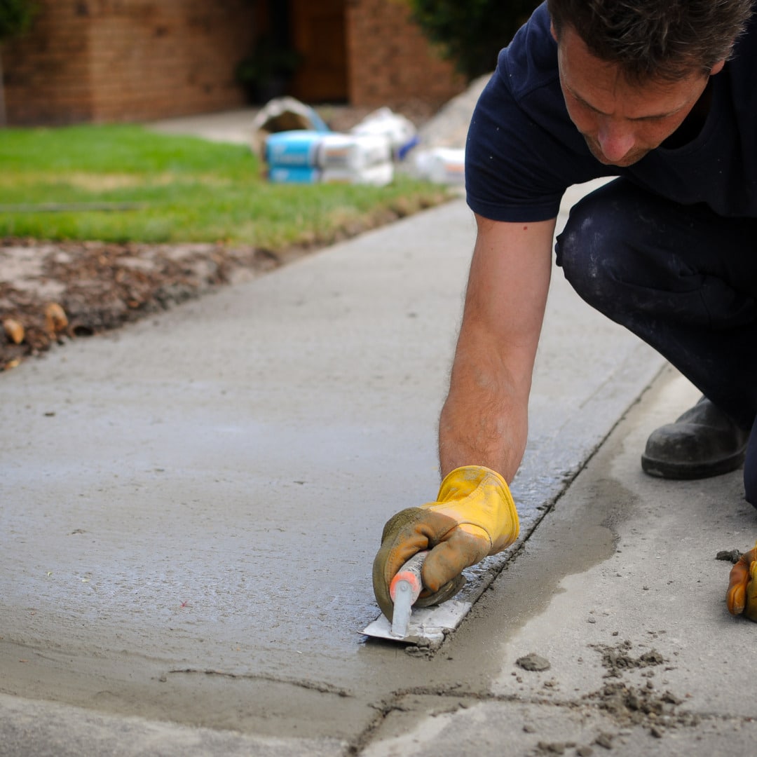 A close-up of a man in a navy shirt and wearing yellow gloves smoothing an edge of a wet concrete sidewalk with a trowel.