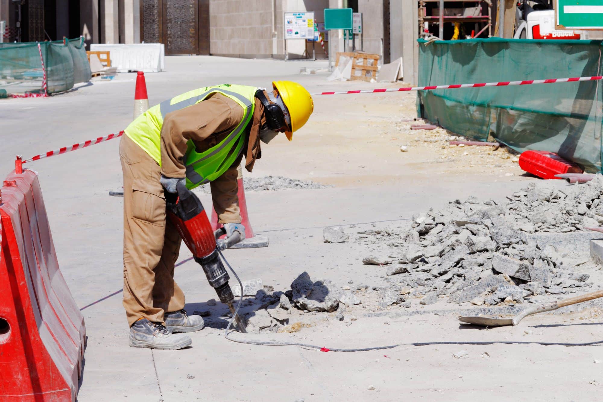 The image depicts a construction worker in a high-visibility vest and yellow hard hat using a red jackhammer to break up concrete on a construction site. There is debris scattered around the worker, indicating active work. The area is cordoned off with red and white safety barriers, and in the background, there's a green construction fence and some temporary site structures. The setting appears to be urban, with a clear sky suggesting a sunny day. The worker is focused on the task, emphasizing safety and work in progress.