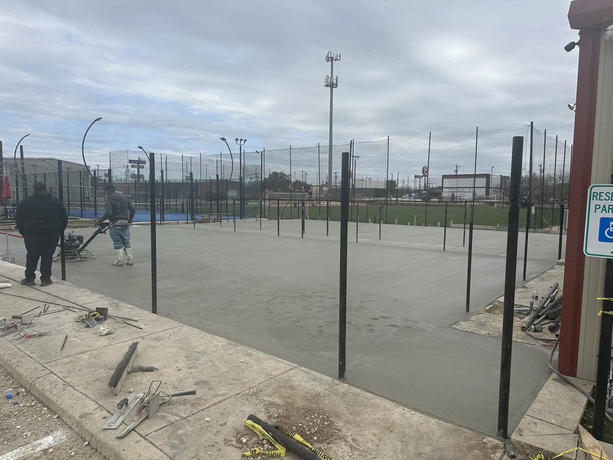Two construction workers are in the process of smoothing a concrete surface, with one operating machinery. The site is framed by metal poles and sports fencing, with tools laid out in the foreground, capturing a moment of development against a backdrop of an overcast day.