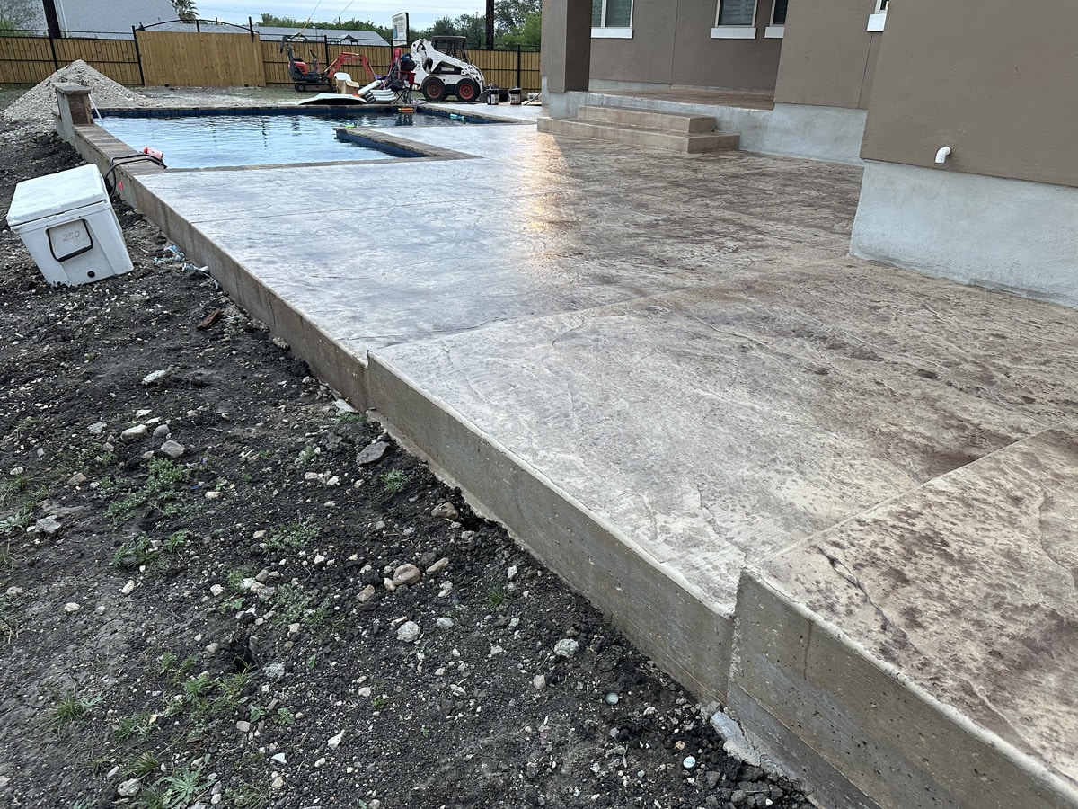 A newly constructed stamped concrete patio is shown beside a tranquil swimming pool, with the unfinished edges and surrounding soil suggesting ongoing backyard landscaping. Construction equipment in the background points to continuous improvement of the leisure area adjacent to the home.