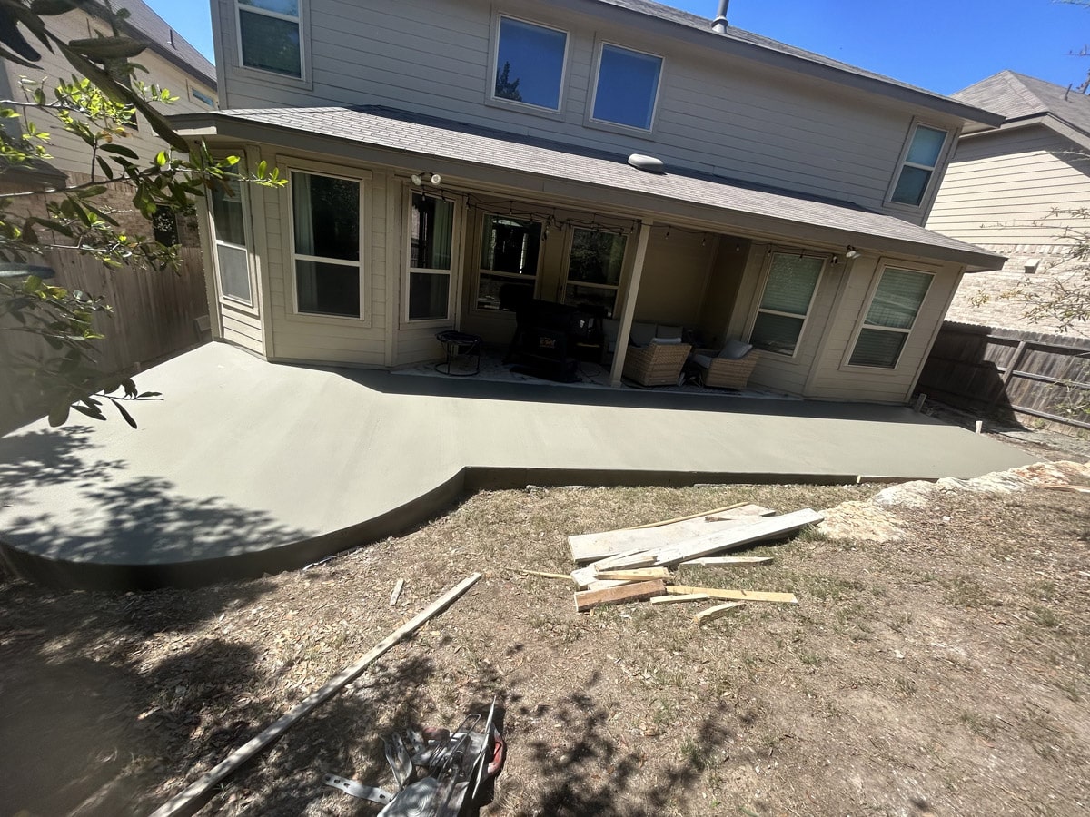 A newly poured, expansive concrete patio curves around the backyard of a suburban home, bordered by a wooden fence and bare soil awaiting landscaping. The patio is bathed in sunlight, with shadows of nearby foliage and construction tools scattered around, indicative of ongoing work.