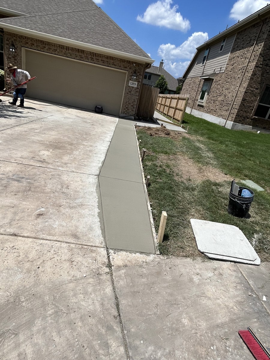 Worker edging a new concrete driveway extension in a suburban San Antonio home with a brick exterior and blue sky overhead.
