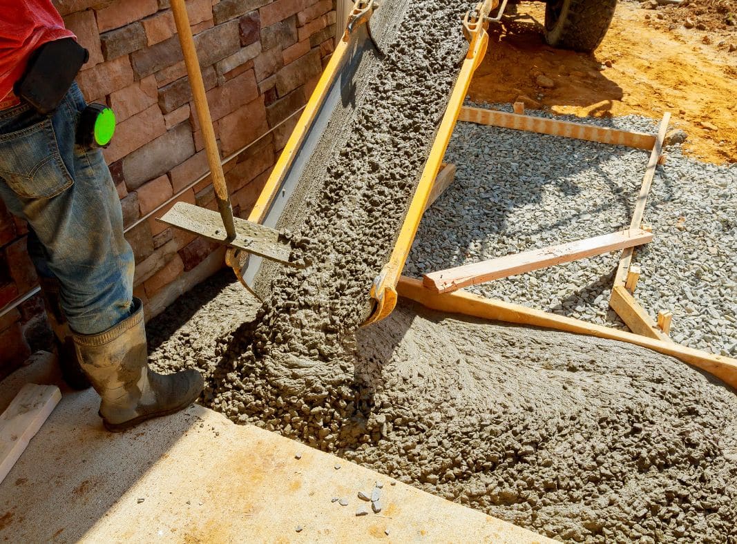 A construction worker in jeans and work boots is using a hoe to spread freshly poured concrete next to a brick wall. The concrete is contained within wooden formwork, indicating the shaping of a foundation or path. The scene is a construction site with visible earth and construction materials, highlighting an active phase of concrete laying.