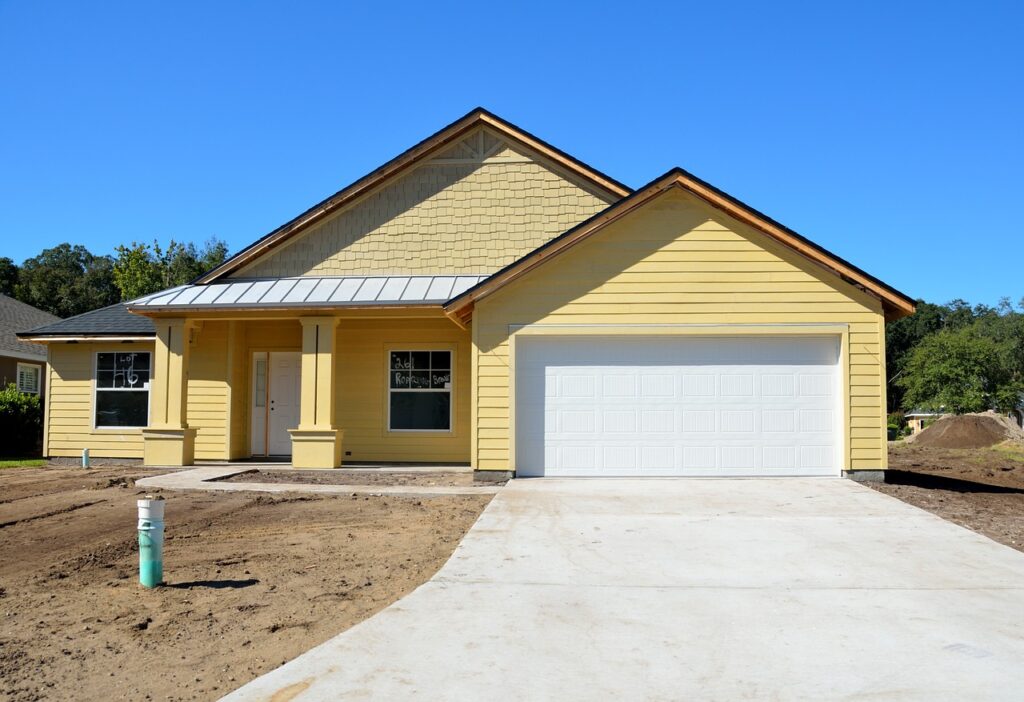 A new single-story home with a light yellow siding and white trim, featuring a large closed white garage door and a covered front entry. The landscaping is unfinished with bare soil and a new concrete driveway.