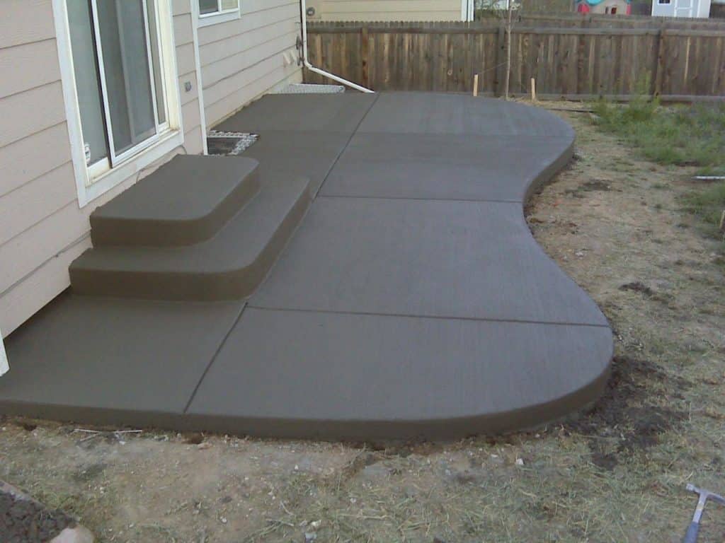 Freshly poured curved concrete patio with steps near a house with a wooden fence.