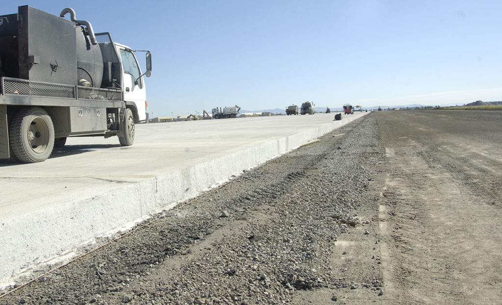 Partially completed concrete road with a sweeper truck on the left and various construction machinery extending into the distance.