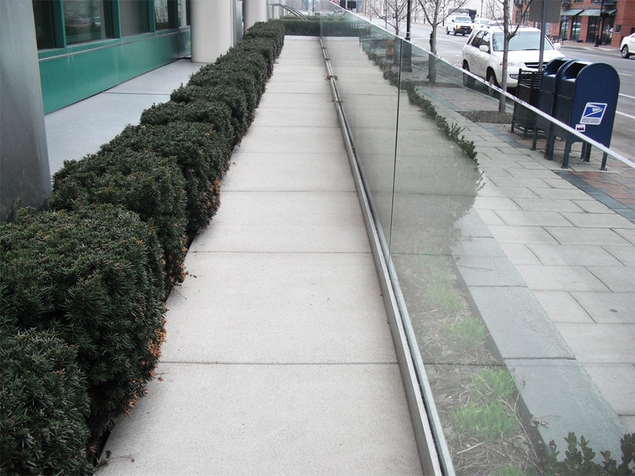 Urban sidewalk with neatly trimmed hedges on the left, a clear glass barrier on the right, a street with parked cars, and a mailbox in the distance.