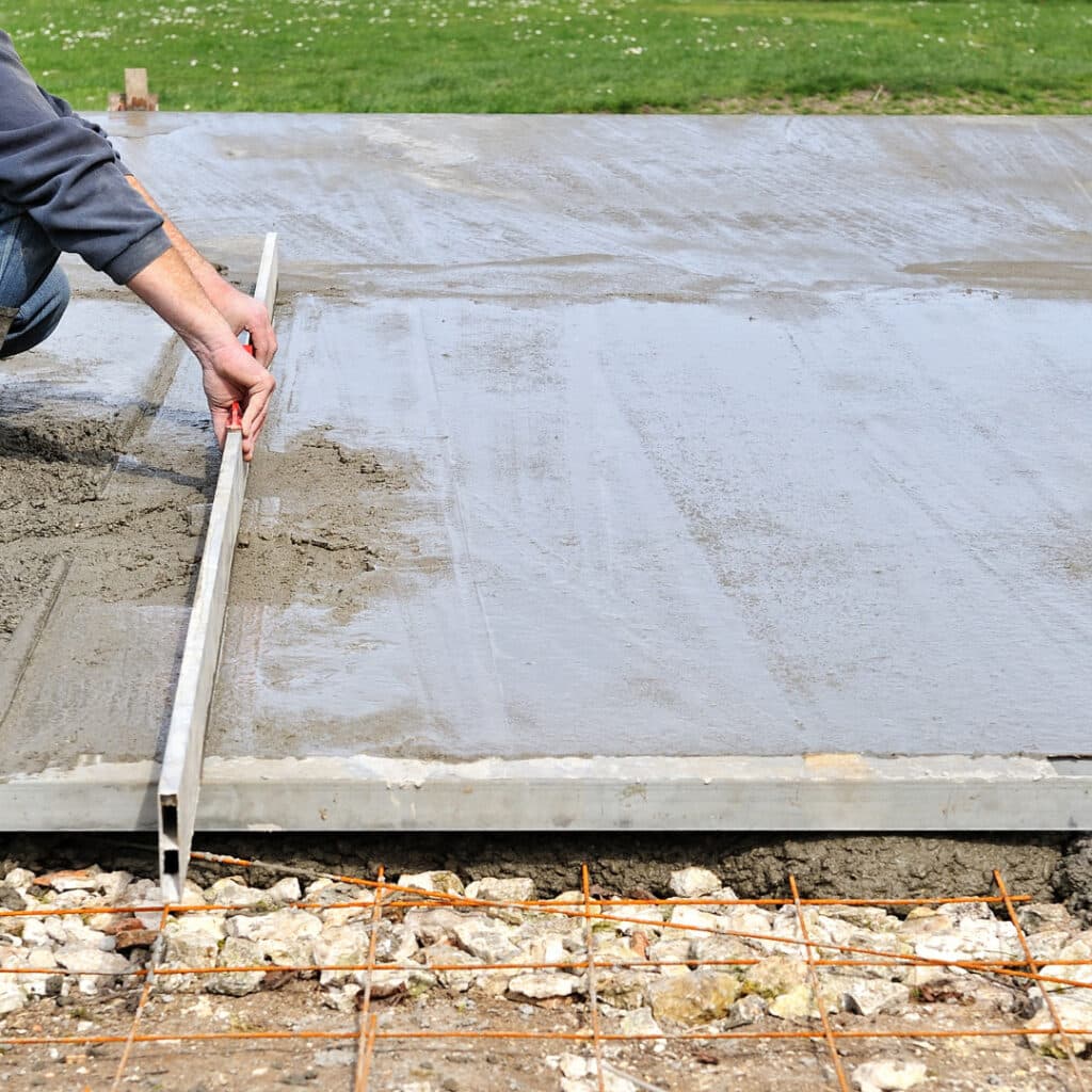 Worker leveling wet concrete on a driveway with a straightedge tool, over a base layer with visible reinforcement mesh.