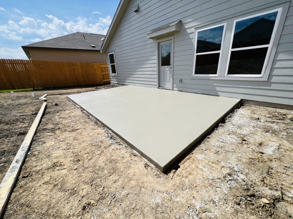 A freshly poured concrete slab in a backyard setting with a clear blue sky above. The slab is adjacent to a light gray house with a wooden fence in the background, reflecting a work in progress with raw earth and construction framing visible.