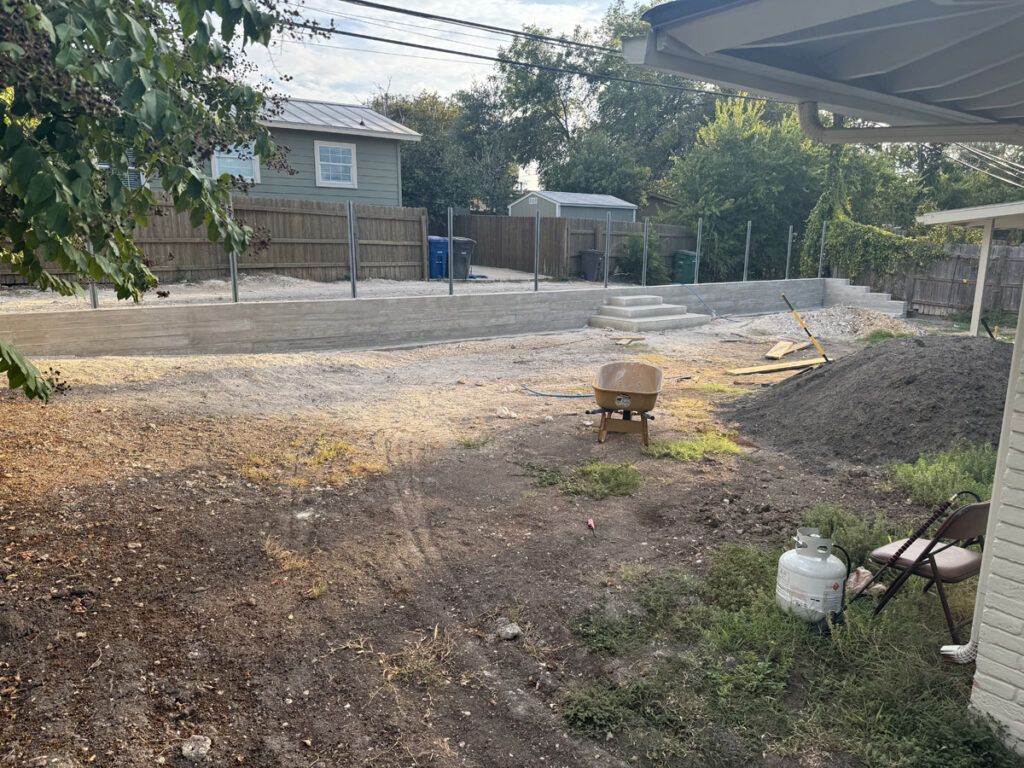 A residential backyard under construction is depicted, with a new concrete wall and stair steps installed at the far end. Metal poles are lined up in preparation for further structure additions. The yard is cluttered with construction materials, including a wheelbarrow, a pile of soil, and a gas canister beside a folding chair. The scene is framed by a green backyard and a wooden fence, with suburban homes in the background, capturing the mix of domestic life and ongoing development.