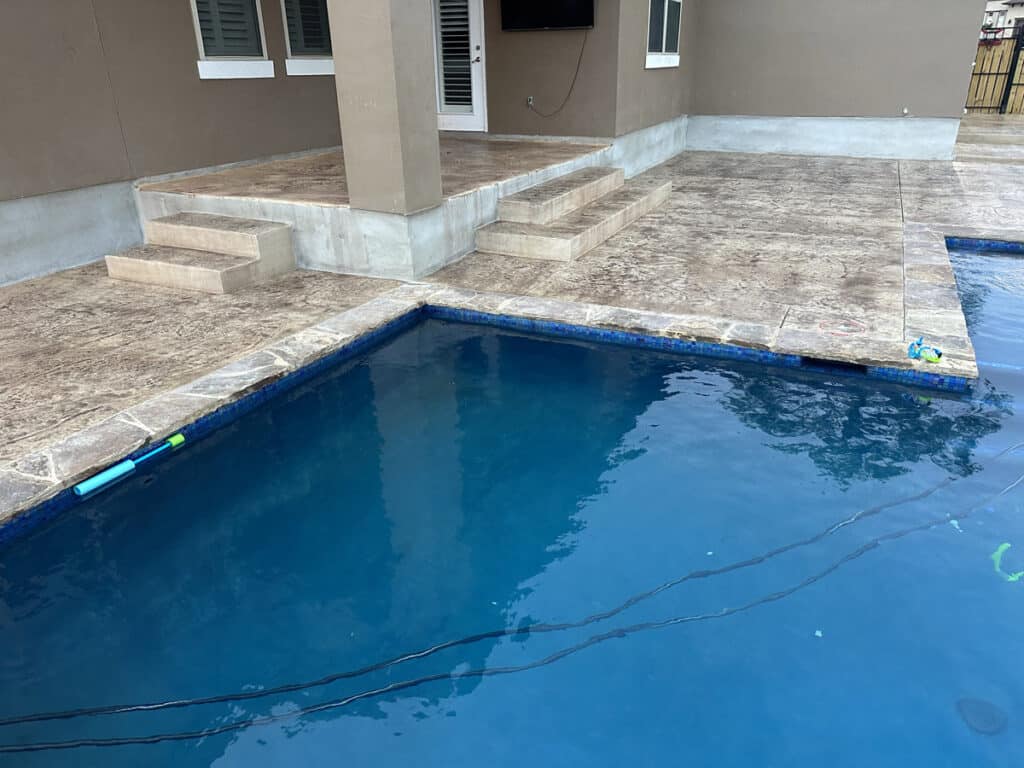 A residential swimming pool with crystal blue water is edged by a newly installed patio with decorative concrete and elegant steps. The pool toys floating in the pool add a playful touch to the serene backyard setting, which is still under final stages of construction.