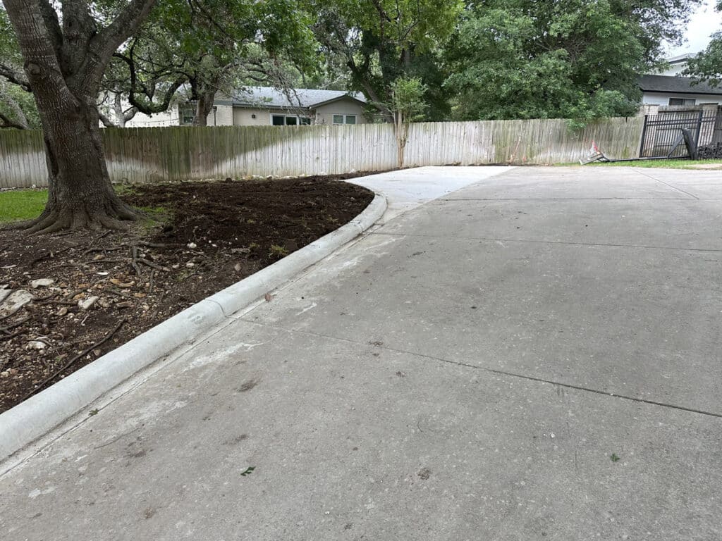 Curved concrete driveway in a residential area with a newly mulched tree bed, wooden fence, and overcast sky.