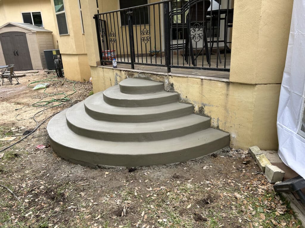 Curved concrete steps recently installed at the side of a yellow building with black metal railings, in the midst of a renovation site in San Antonio, Texas. Visible are the building’s exterior details, renovation equipment, and a partially seen white tent, indicative of ongoing construction.