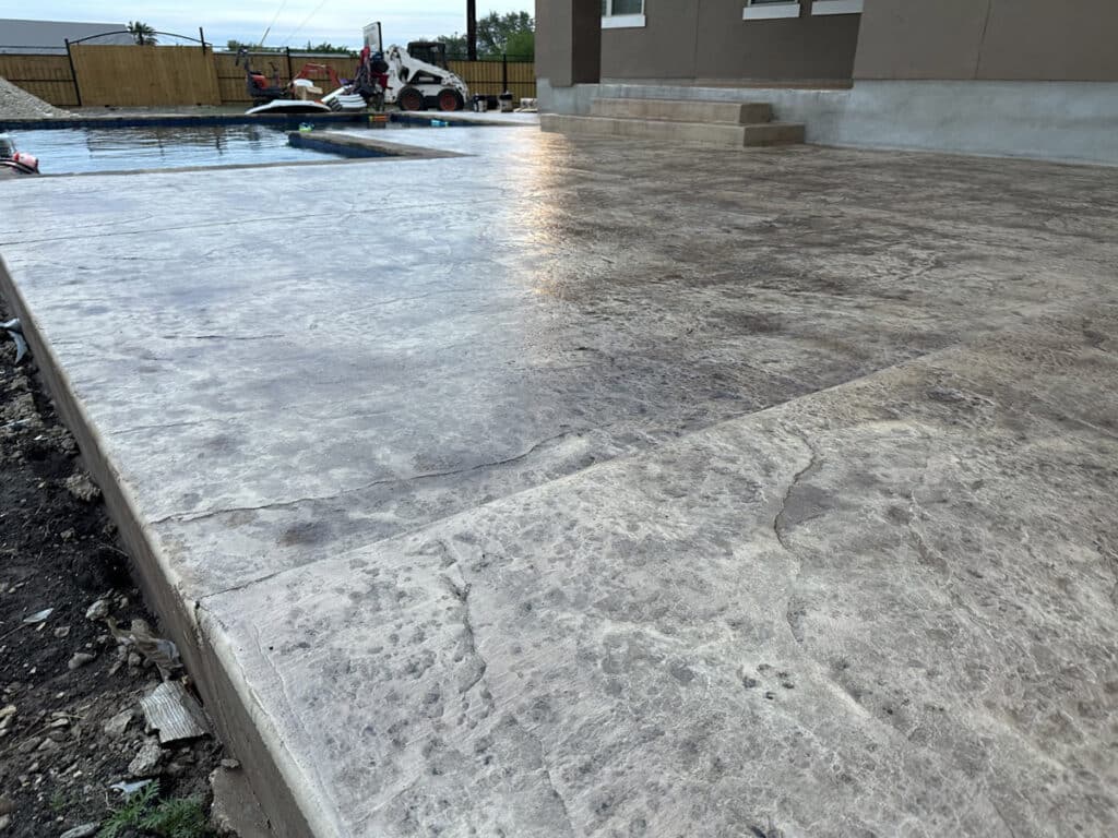 A newly poured and textured concrete patio glistens with moisture, with construction equipment and a building in the background.