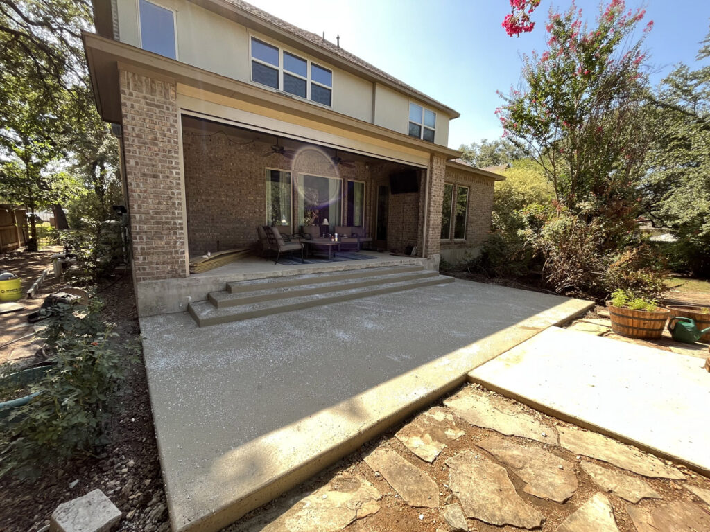 A freshly poured concrete patio extension dries in the sunlight, adjoining an existing stone patio outside a two-story brick home. The extension includes a set of three concrete steps leading to the yard. The house is partially shaded by a mature tree with bright pink blossoms, and the landscaped garden includes potted plants and shrubbery. The scene is a blend of construction and cozy domesticity, with outdoor furniture and a lawn mower in the background, suggesting ongoing home improvement efforts.