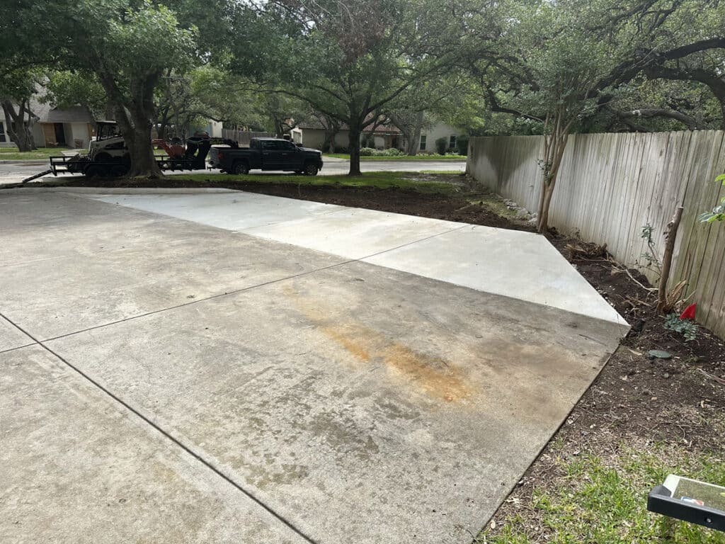 A concrete driveway expansion is featured in a residential area, with a clear demarcation between the new and older sections. Vehicles and a trailer parked under the shade of a large tree suggest a home improvement project in progress, with a wooden fence and homes in the background providing a suburban context.