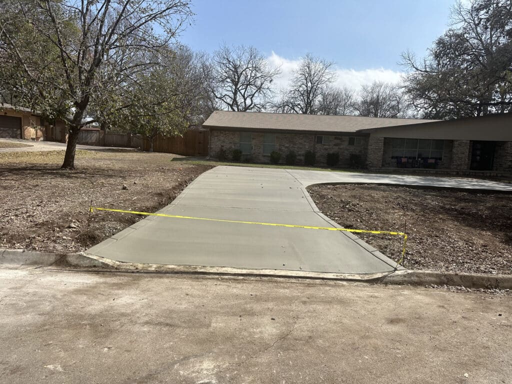 A newly poured and smoothed concrete driveway in front of a residential house is sectioned off with yellow caution tape. The driveway, bordered by a bare winter lawn and under the shade of a leafless tree, awaits its first use on a sunny day.