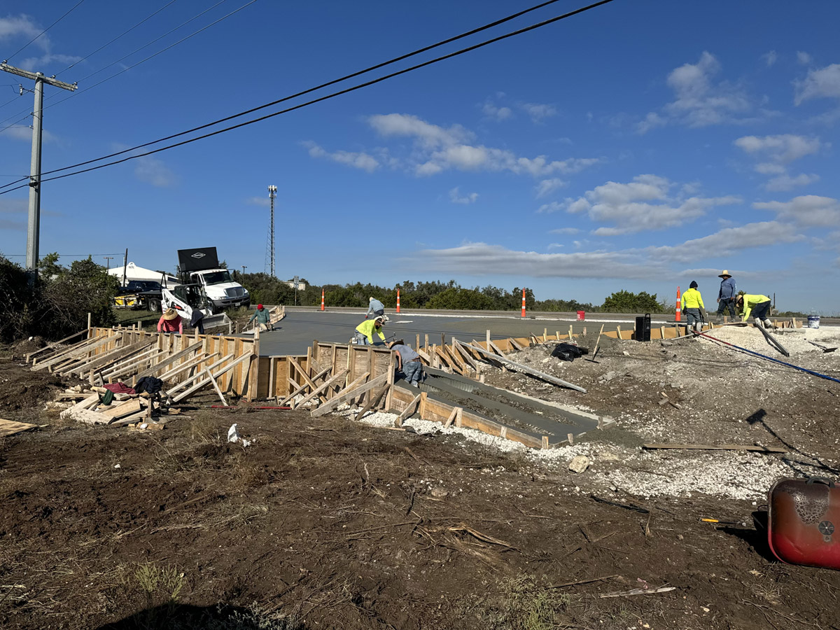 Active construction scene with workers in high-visibility clothing engaged in concrete work beside a busy road. A cement truck is on standby as workers spread and smooth freshly poured concrete. The construction area is bordered by orange safety cones and wooden formwork. A clear sky with scattered clouds looms above the scene, while power lines and poles stand prominently in the background, suggesting an infrastructure development in a suburban area.