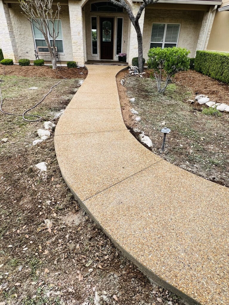 A newly installed pebble-textured pathway leads to the entrance of a suburban home, flanked by manicured shrubs and a well-maintained lawn. The residence features a stone facade and a welcoming front door, with a garden hose laid out to the side, indicating recent landscaping work.