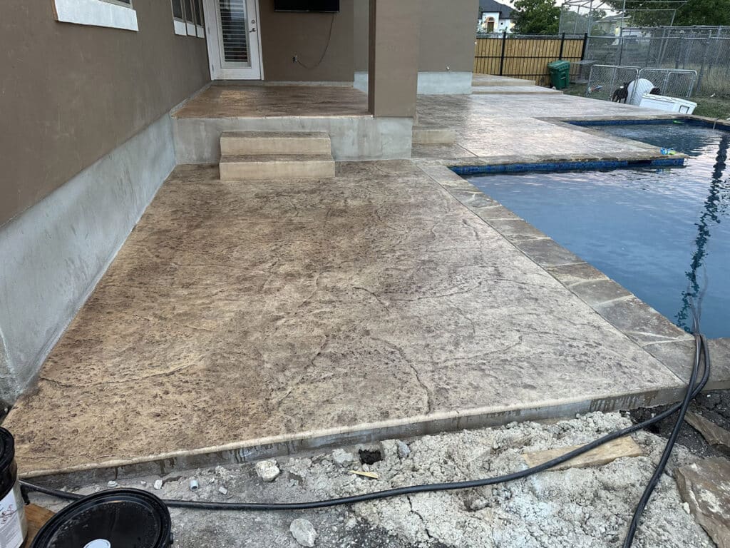 The image shows a newly constructed patio with textured concrete and steps leading up to a house, complementing the adjacent swimming pool. Visible construction materials and a hose across the scene indicate ongoing outdoor improvements and landscaping work.