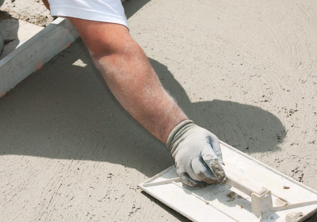 Close-up of a construction worker's arm using a hand float to smooth wet concrete, with formwork visible in the background.