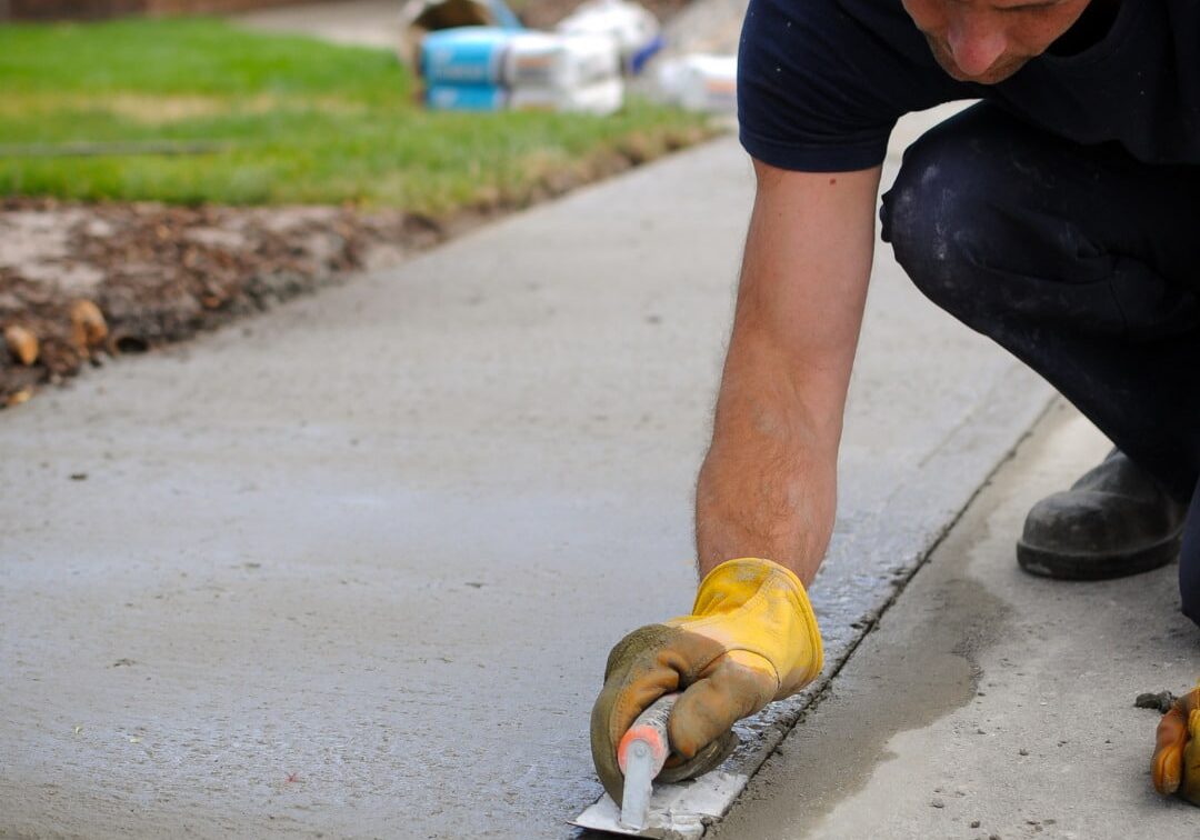 A close-up of a man in a navy shirt and wearing yellow gloves smoothing an edge of a wet concrete sidewalk with a trowel.