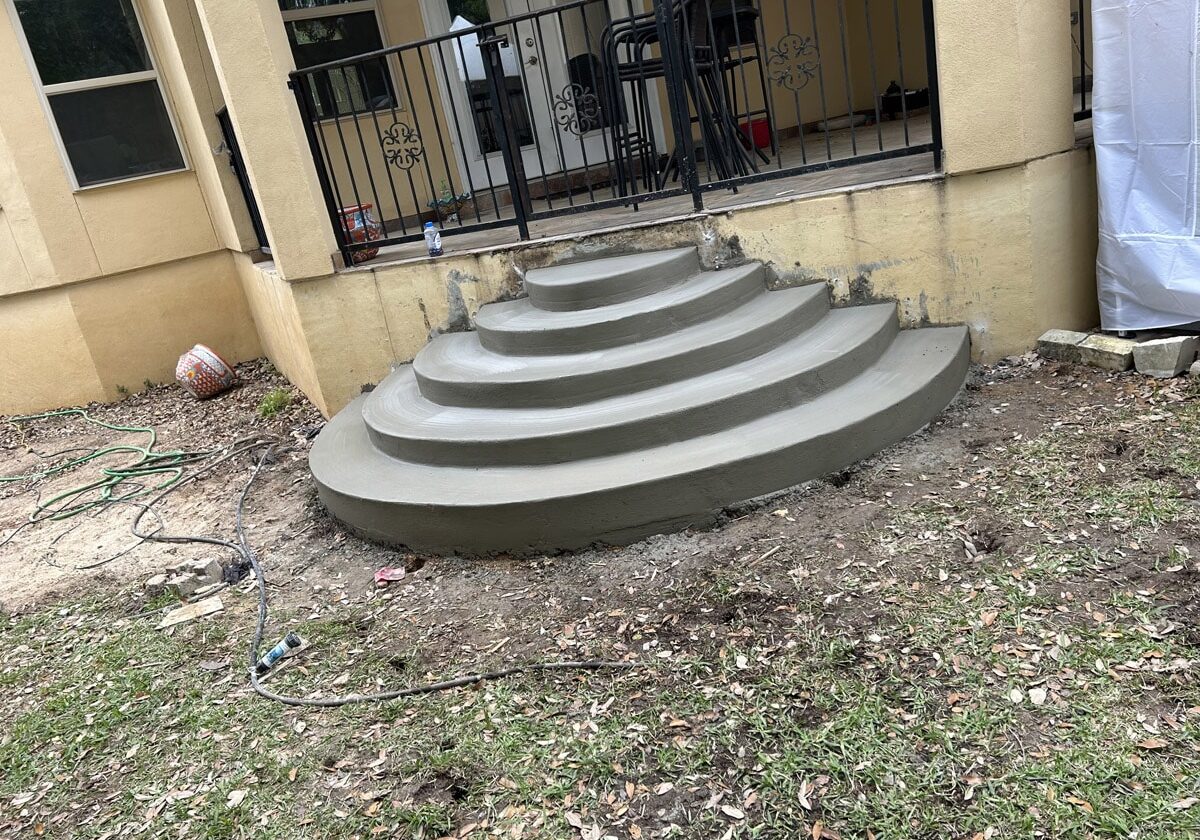 Architectural detail of newly constructed, arching concrete steps in front of a building with a yellow facade and decorative black railings in San Antonio, Texas. The surrounding area is strewn with construction debris, a green garden hose, and a white plastic covering, indicating ongoing outdoor work.