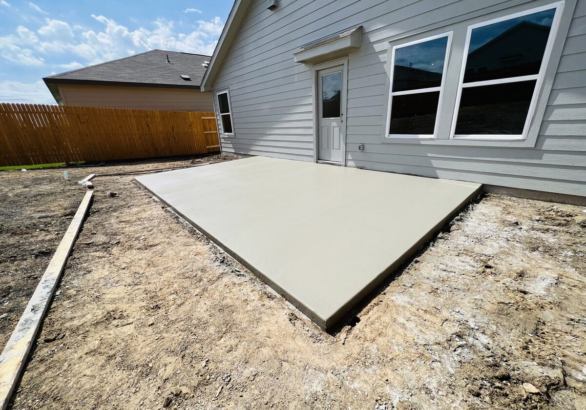 A freshly poured concrete slab in a backyard setting with a clear blue sky above. The slab is adjacent to a light gray house with a wooden fence in the background, reflecting a work in progress with raw earth and construction framing visible.
