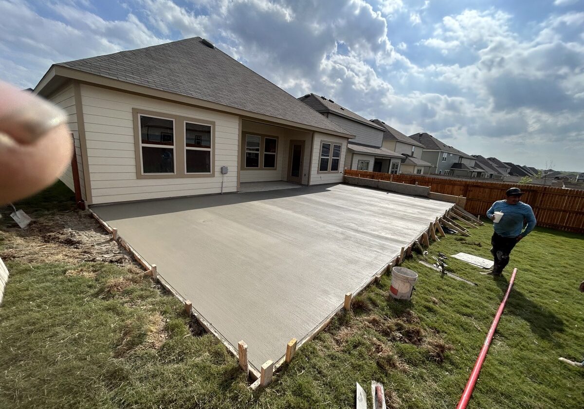 Large backyard patio extension freshly poured and smoothed, edged with wooden framing, with a construction worker carrying equipment on the lush green lawn. The suburban home and the expansive new patio are illuminated by the afternoon sun under a dramatic sky.