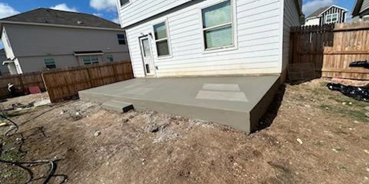 Freshly poured concrete patio extension at the back of a suburban home, with the backyard area under construction and surrounding wooden fencing.
