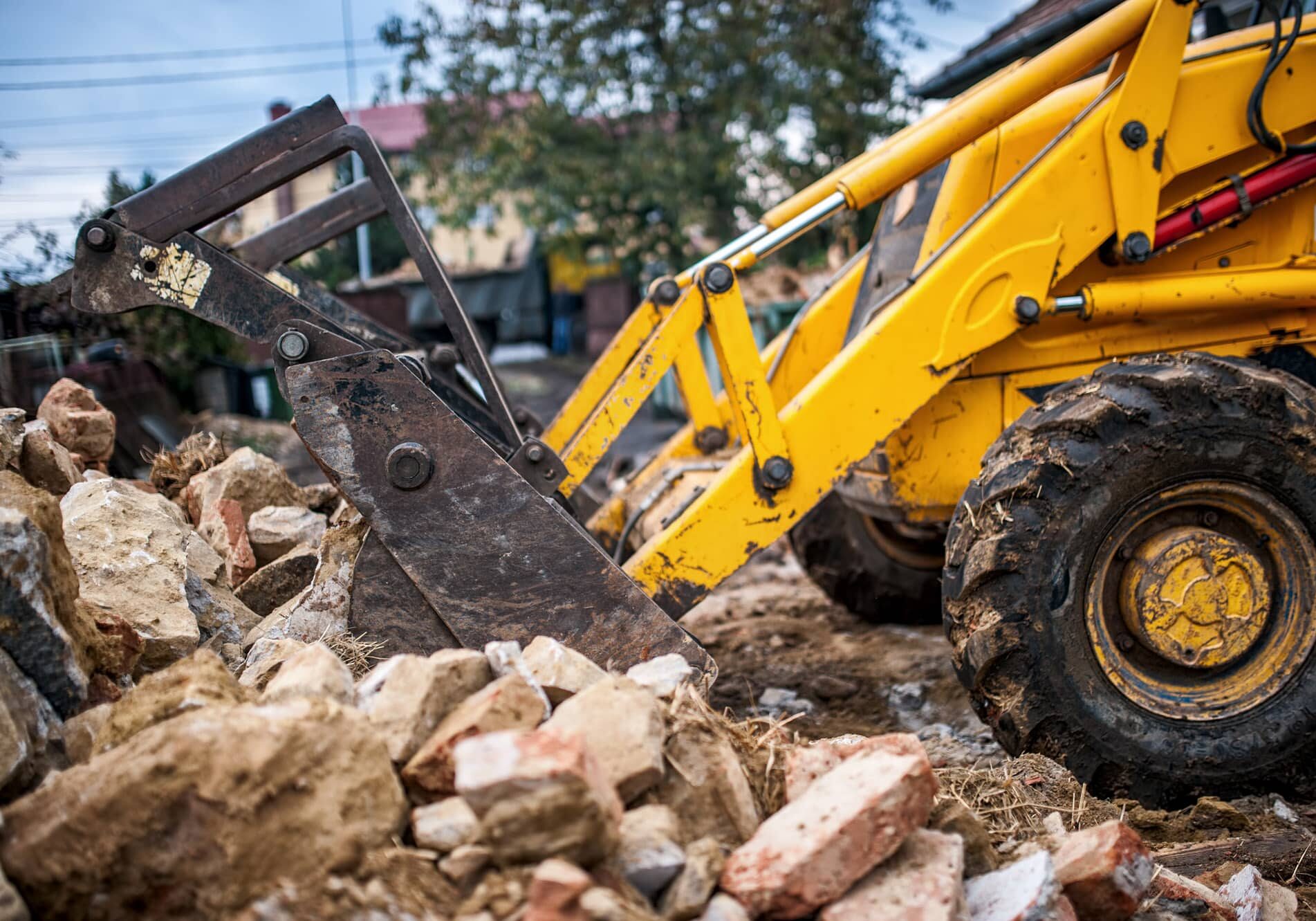 Yellow construction backhoe with extended bucket among rubble and debris at a demolition site.