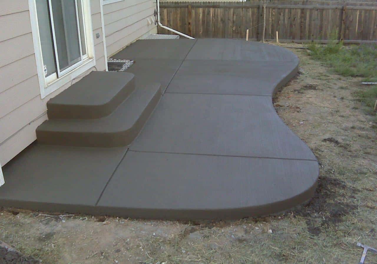 Freshly poured curved concrete patio with steps near a house with a wooden fence.