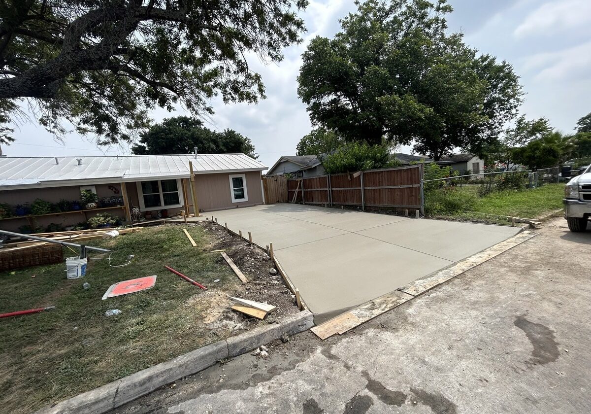 Freshly poured concrete driveway extension in front of a single-story home with a solar panel roof. The driveway is bordered by wooden forms and a large tree provides shade overhead. Construction materials and tools are visible on the grassy area adjacent to the driveway.