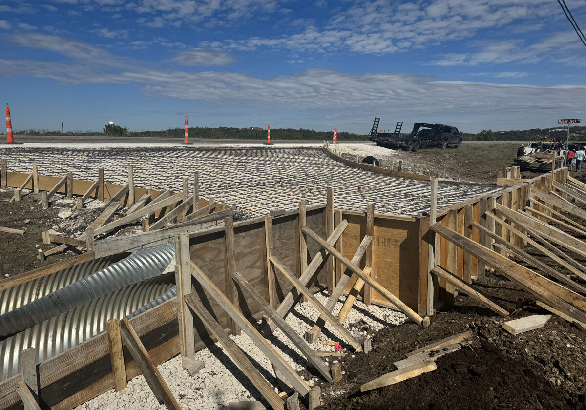 A construction site under clear blue skies showcasing the foundation work for a new road or infrastructure project. Safety cones mark the perimeter where a concrete base has been laid out, featuring a grid of reinforcing steel bars ready for a concrete pour. Construction vehicles, including a black truck and heavy machinery, are parked on the adjacent unpaved land. The scene is busy with construction activity, materials, and equipment, with a backdrop of open skies and distant trees.