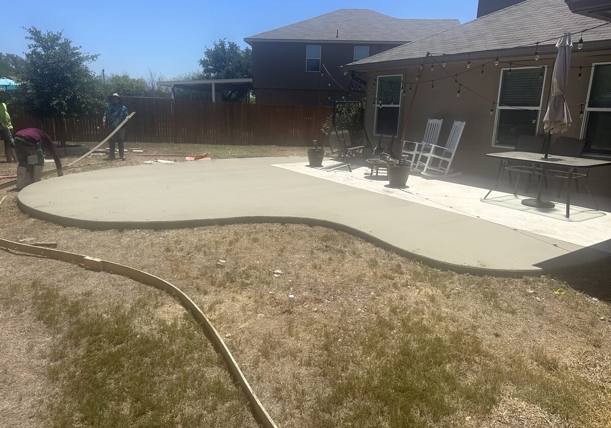 Construction workers finishing a large, curved concrete patio in a sunny backyard with seating area and green grass, in San Antonio.