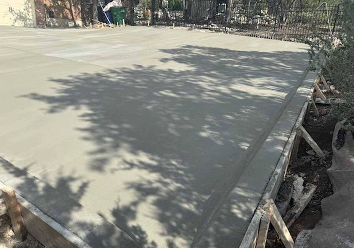 A broad expanse of freshly laid concrete driveway is shaded by the intricate patterns of tree shadows, with construction frameworks still visible along the edge. The scene captures the contrast between the ruggedness of the wooden forms and the smooth finish of the driveway, set against a residential backdrop.