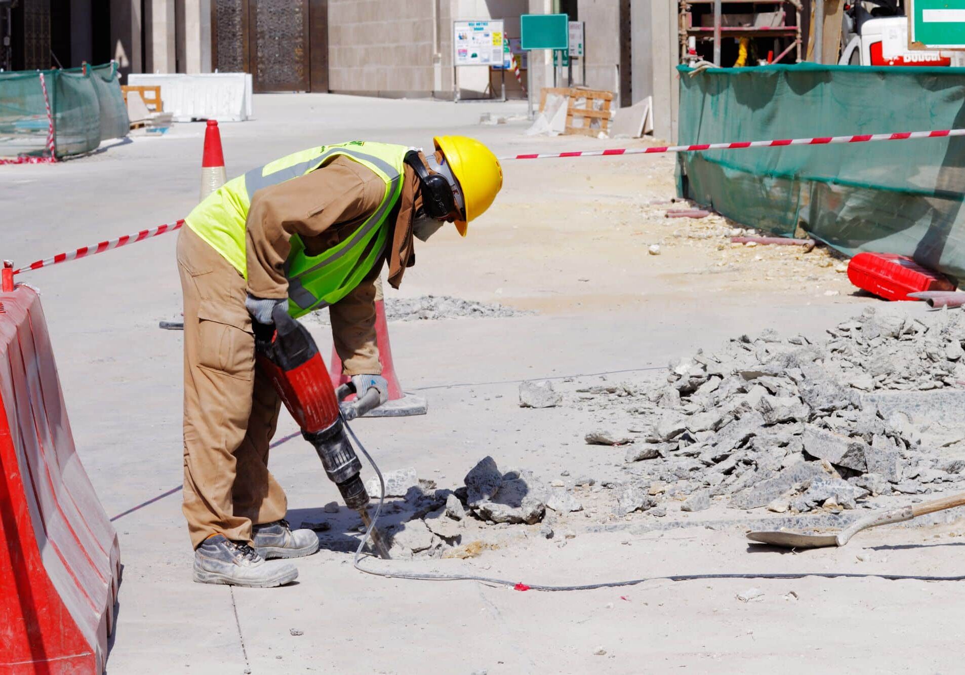 The image depicts a construction worker in a high-visibility vest and yellow hard hat using a red jackhammer to break up concrete on a construction site. There is debris scattered around the worker, indicating active work. The area is cordoned off with red and white safety barriers, and in the background, there's a green construction fence and some temporary site structures. The setting appears to be urban, with a clear sky suggesting a sunny day. The worker is focused on the task, emphasizing safety and work in progress.