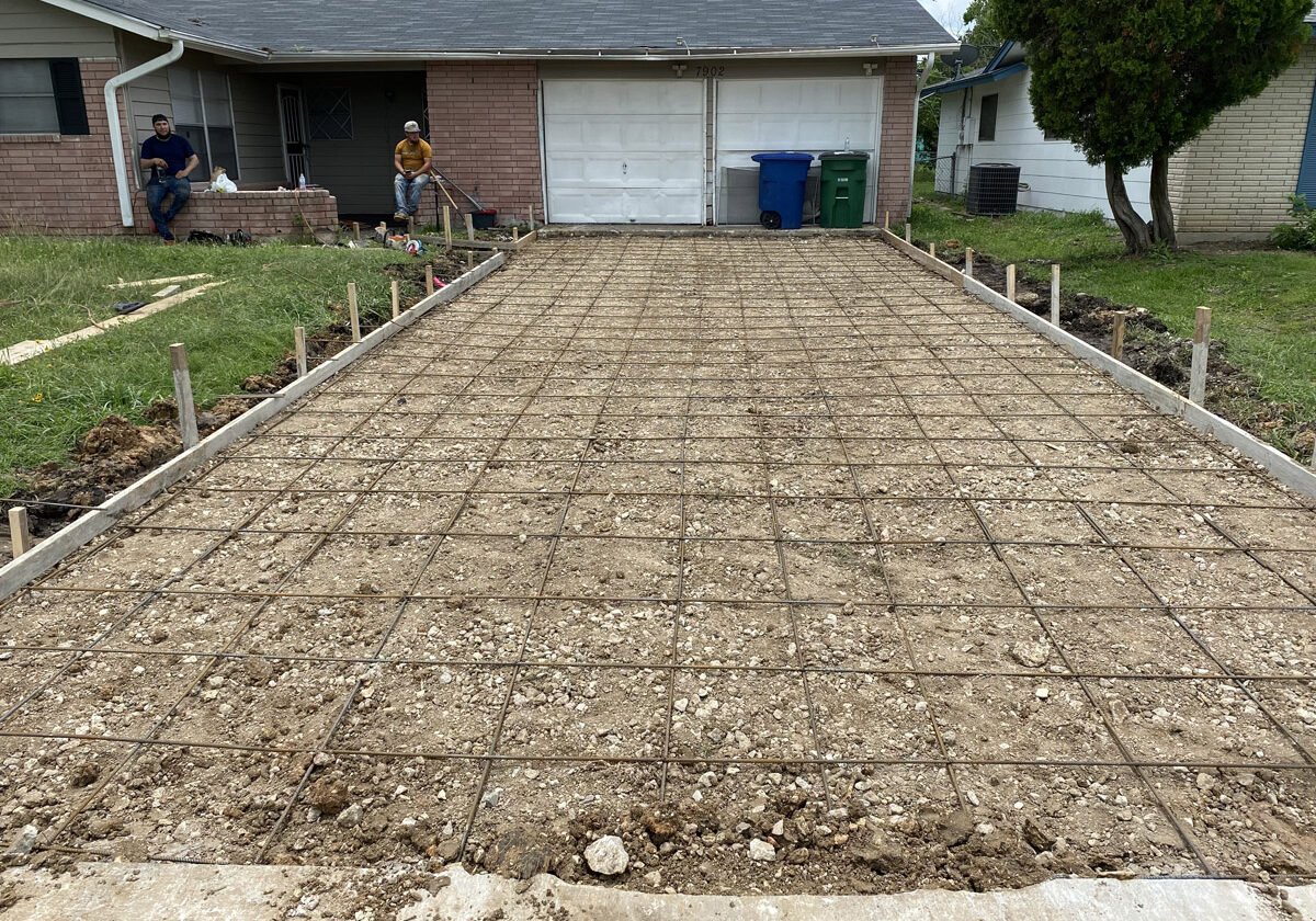 A residential driveway-to-be is laid out with a grid of rebar, ready for concrete pouring, framed by wooden forms. Two workers rest in the background near the home's entrance, suggesting a brief respite in a day of hard work under an overcast sky.