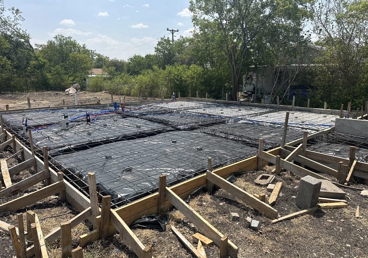 Construction site in San Antonio with a large area prepared for concrete foundation work, featuring steel rebar grid patterns. Wooden formwork creates sections for pouring concrete. Trees and a house are visible in the distance, under a sunny Texas sky.