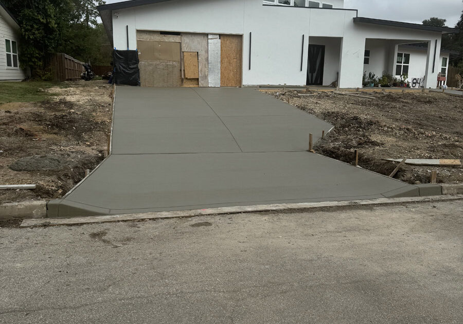 A freshly poured concrete driveway leading to a modern house under construction. The driveway is divided into sections by control joints, ensuring proper expansion and contraction. The house, partially obscured by protective materials, features a contemporary design with a flat roof and geometric lines. A stark contrast is visible between the smooth, grey surface of the concrete and the rough, unfinished landscape around. The overcast sky suggests a recent completion of the concrete work, waiting to cure.