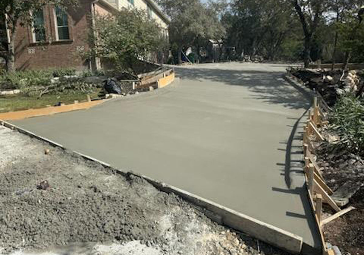 Freshly poured concrete forms a new driveway section next to a two-story brick house. The driveway is neatly bordered by wooden forms, and the adjacent lawn is dotted with cut branches and debris, indicative of recent landscaping. Mature trees provide shade over the area, and the background reveals a quiet residential street lined with similar houses, under a clear sky with ample daylight.