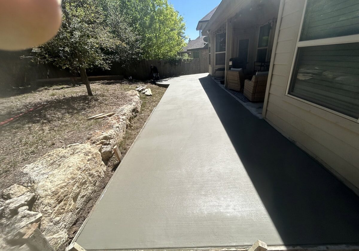A newly constructed smooth concrete walkway stretches alongside a residential home, flanked by a natural rock edge and a neatly trimmed yard. The bright sunlight casts sharp shadows, highlighting the clean lines of the walkway still bordered by wooden forms.