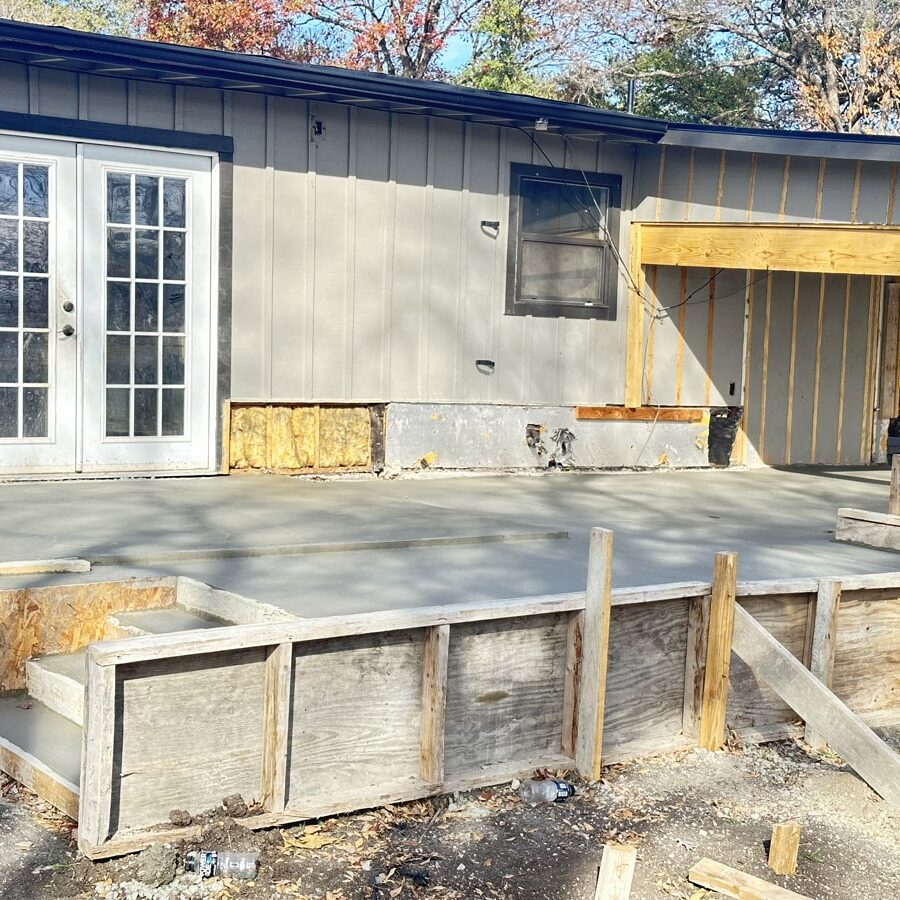 A newly poured concrete foundation sits adjacent to a house undergoing renovations. French doors and a partially insulated exterior wall are visible, with the foundation framed by wooden supports. The clear shadows and bright sunlight indicate work in progress on a crisp day.