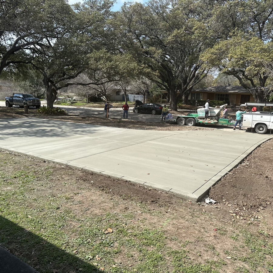 Workers are finalizing a new concrete driveway in a residential area shaded by large oak trees. The scene captures the workers with their equipment and vehicles, set against a backdrop of suburban homes on a sunny day, symbolizing neighborhood development.