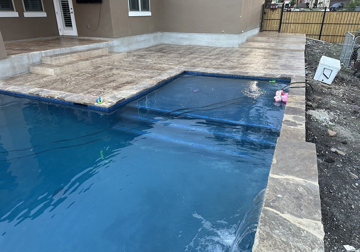 An inviting swimming pool with clear blue water is bordered by a newly constructed patio with textured concrete and steps leading to the home's backdoor. Children's pool toys float on the water, hinting at family fun, amidst a yard still under renovation.
