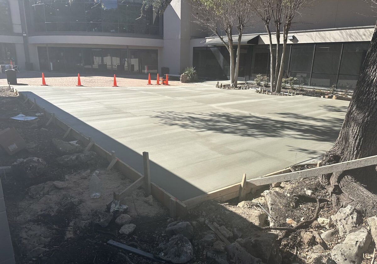 Parking lot paving Freshly poured concrete courtyard with orange cones and wooden formwork, near a commercial building and trees casting shadows.