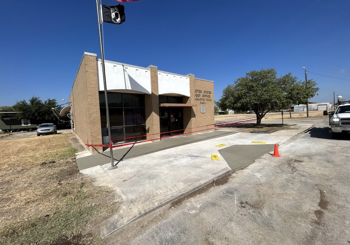 Exterior of a United States Post Office with a newly constructed concrete access ramp and tactile paving, cordoned off with red tape, on a sunny day in San Antonio.